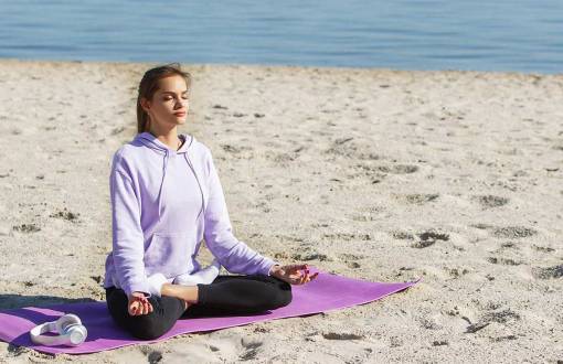 Eine junge Frau in violetten Sportklamotten sitzt am Strand und macht Yoga im Ostseeurlaub in den Arkona Strandresidenzen.