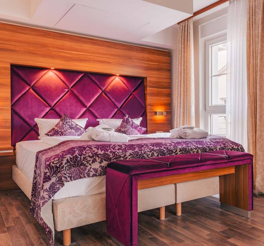 Doppelbett im Suite Apartment der Arkona Strandresidenzen Binz mit violettem Kopfteil und eleganter Bettbank vor hellem Fenster