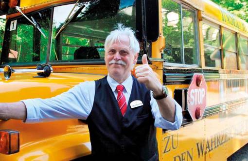 Ein Mann in Uniform lächelt in die Kamera und steht vor einem gelben Schulbus bereit für einen Ausflug der Arkona Strandresidenzen.